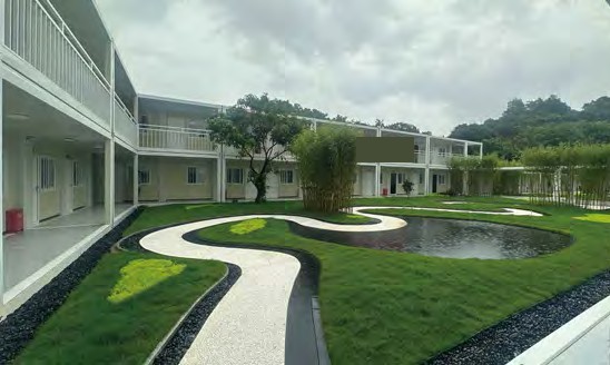 Two-storey modular project office buildings enclosing a landscaped courtyard with a curved paved path, lawn, bamboo planting and a water feature